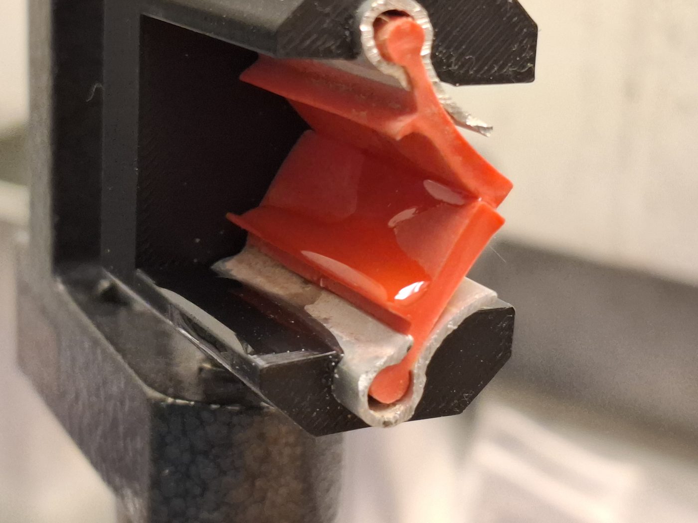 Macro photograph of the scraper under test: a red rubber blade seated in its holder, wet with epoxy, with carbon fibre tow drawn through the gap beneath it